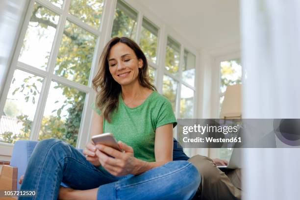 smilong mature woman sitting on couch at home using cell phone with man in background - ehefrau stock-fotos und bilder