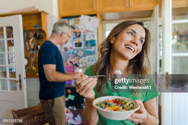 happy mature woman at home eating a salad with man in background - man eating woman out ストックフォトと画像