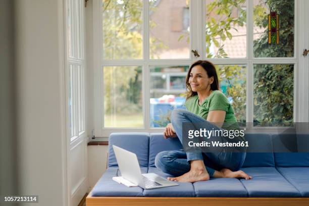 smiling mature woman sitting on couch at home with laptop - older woman with brown hair foto e immagini stock