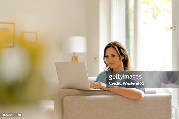 mature woman sitting on couch at home using laptop - notebook stockfoto's en -beelden