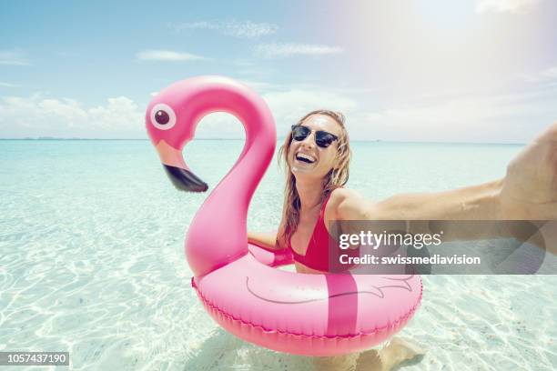 young woman takes selfie portrait on idyllic beach with inflatable pink flamingo in pristine clear water in the islands of thailand. people travel luxury fun and cool attitude concept - tube imagens e fotografias de stock