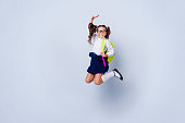 Funky nice cute genius positive cheerful lovely small little girl with curly pigtails in white formal blouse shirt, skirt, jumping, raising hand up, fooling, yellow bag. Isolated over grey background