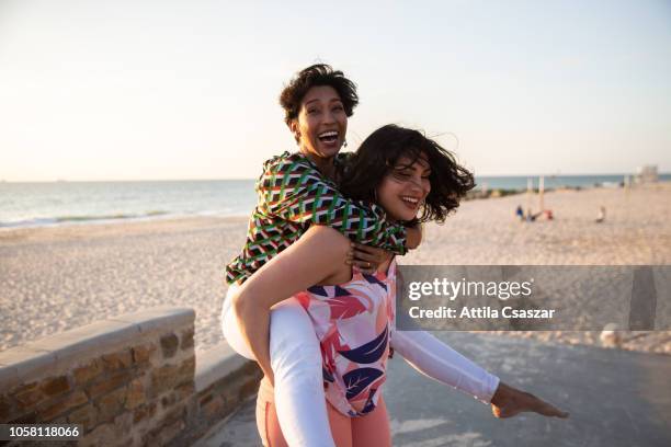 young women piggybacking on sandy beach at sunset - life events foto e immagini stock