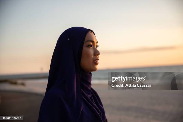 young woman wearing hijab looking at distance at sunset at beach - muslim woman beach stock pictures, royalty-free photos & images