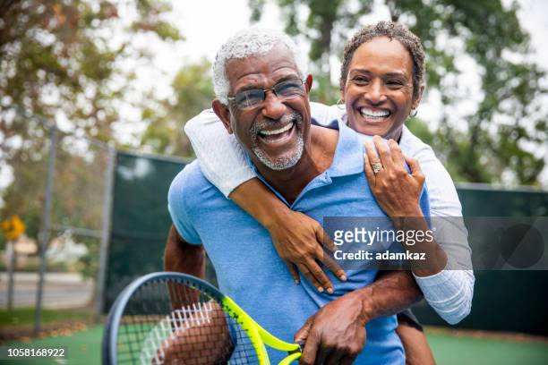 coppia nera senior sul piggyback del campo da tennis - vita attiva foto e immagini stock