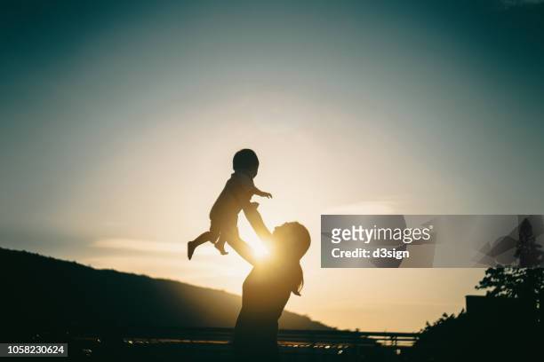 silhouette of mother raising baby girl in the air outdoors against sky during a beautiful sunset - baby carrier outside bildbanksfoton och bilder