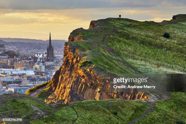 rocky cliffs of salisbury crags in holyrood park with edinburgh city the in background at sunset - edimburgo foto e immagini stock