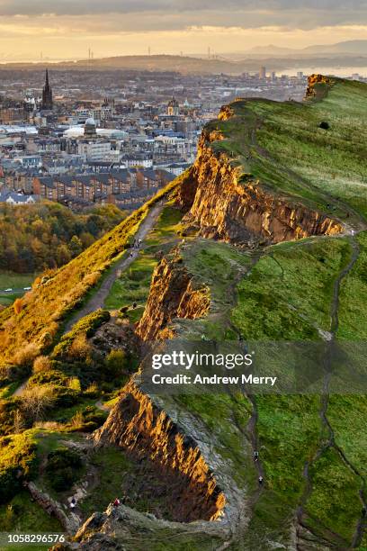 rocky cliffs of salisbury crags in holyrood park with edinburgh city the in background at sunset - holyrood park stock-fotos und bilder