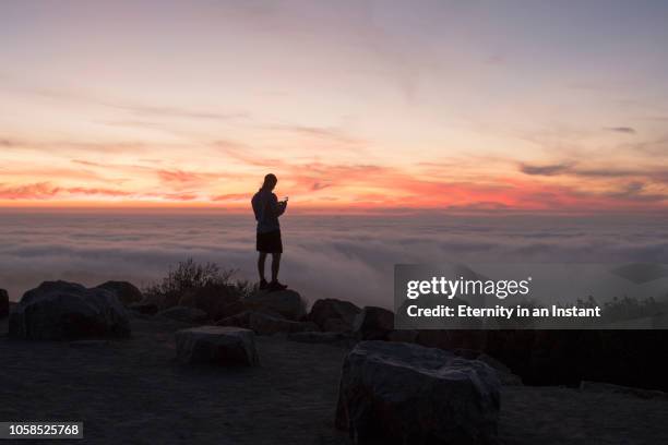 young man standing on a mountain top at sunset - back lit clouds stock pictures, royalty-free photos & images