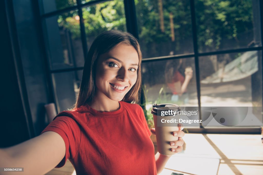 Hermosa joven haciendo selfie en la cámara frontal de su smartphone y sosteniendo una taza de papel con una bebida