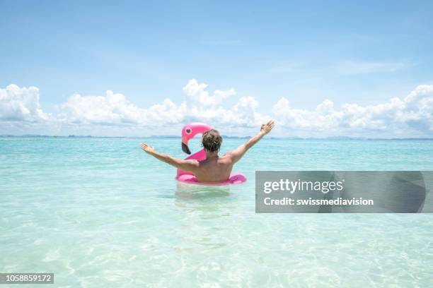 freudiger junger mann schwimmende mit aufblasbarer flamingo an wunderschönen tropischen strand strand urlaub im paradies. männlich outstretches arme für glück und vitalität - glory tube stock-fotos und bilder