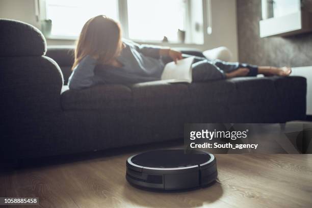 woman reading book while robotic vacuum cleaner cleaning house - robot vacuum stock pictures, royalty-free photos & images