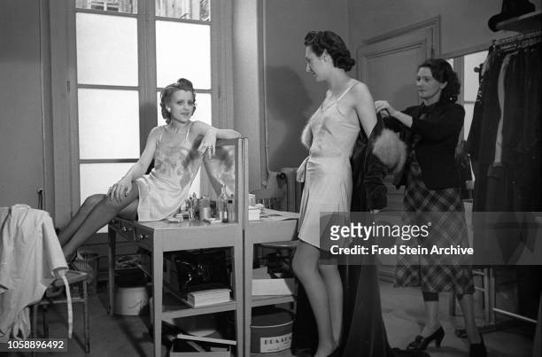 As an unidentified model in a slip sits on a make-up table, a second model is helped into a floor-length, fur-trimmed, velvet gown, Paris, France,...