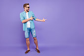 Full body size length of attractive bearded trendy stylish confident cheerful man, wearing denim and glasses, pointing with fingers aside, over bright violet purple background, empty blank copy space