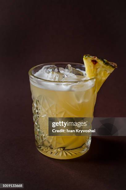 glass of yellow alcohol cocktail with ice and slice of pineapple on elegant dark brown background - cocktail isolated stock-fotos und bilder