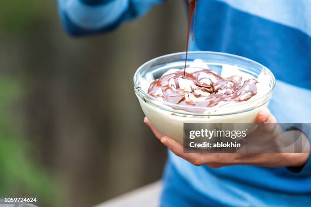 Closeup of young man outside, outdoors, holding glass bowl of homemade, raw vegan vanilla ice cream, adding chocolate liquid sauce from spoon