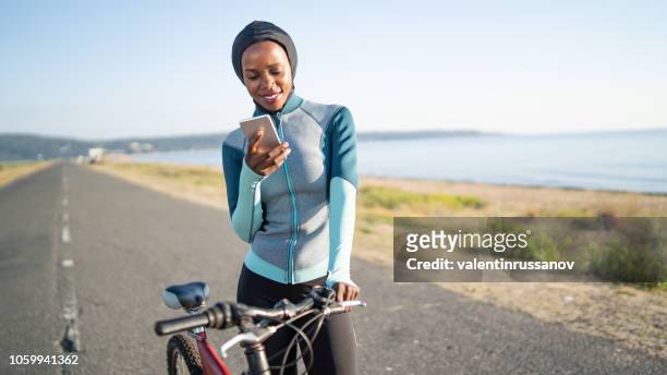 african sportswoman with hijab, with a bike using smart phone on road - remote location stock pictures, royalty-free photos & images