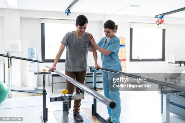 concentrado de paciente do sexo masculino na fisioterapia, andando com a ajuda de barras paralelas e o terapeuta ao lado dele - physical injury - fotografias e filmes do acervo
