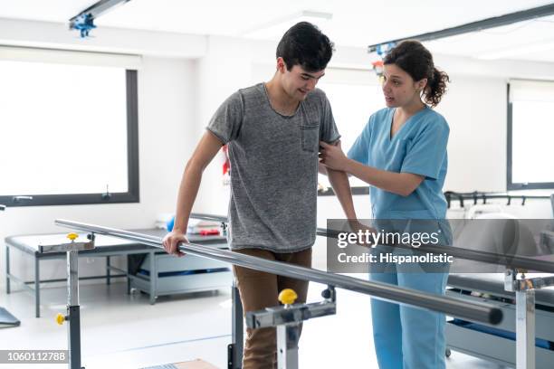 young male patient leaning on parallel bars to walk while female therapist is motivating him - parallel bars stock pictures, royalty-free photos & images