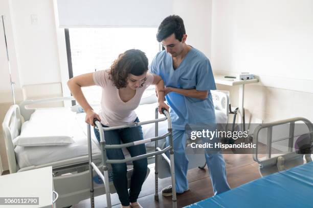 verpleger te helpen een jonge patiënt stand up van ziekenhuisbed terwijl ze op walker leunt - injured man in hospital bed stockfoto's en -beelden