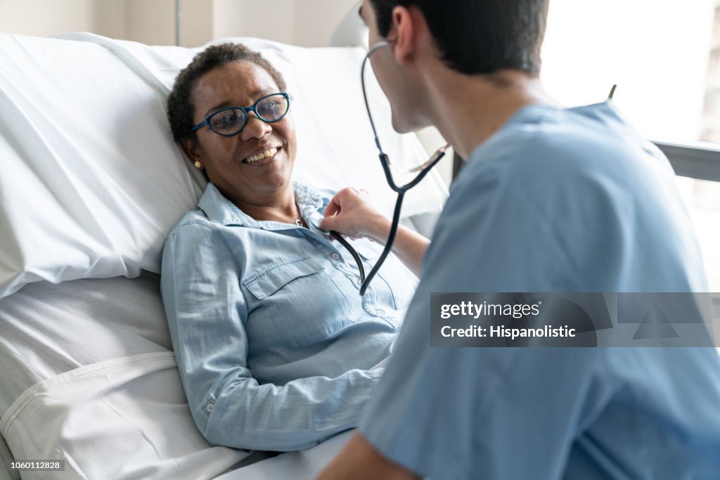 Female black patient lying down on hospital bed while nurse is checking her heart beat with stethoscope