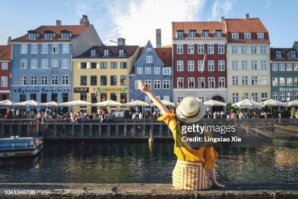young woman sitting in front of colorful buildings along nyhavn (new harbour), copenhagen, denmark - copenhagen foto e immagini stock