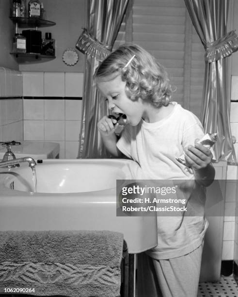 1950s LITTLE BLOND GIRL IN PAJAMAS STANDING AT BATHROOM SINK BRUSHING HER TEETH TOOTHPASTE AND TOOTHBRUSH IN HAND