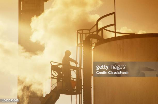 1990s MAN ANONYMOUS REFINERY WORKER ON LIFT OUTSIDE STORAGE TANK SILHOUETTED AGAINST SMOKE