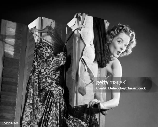 1950s FLIRTING WOMAN LOOKING AT CAMERA PEEKING AROUND FROM BEHIND DRESSING SCREEN HUNG WITH DRESS BRA AND SLIP