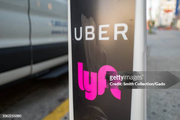 Close-up of vertical sign with logos for ridesharing companies Uber and Lyft, with wheels of a car in the background, indicating a location where...
