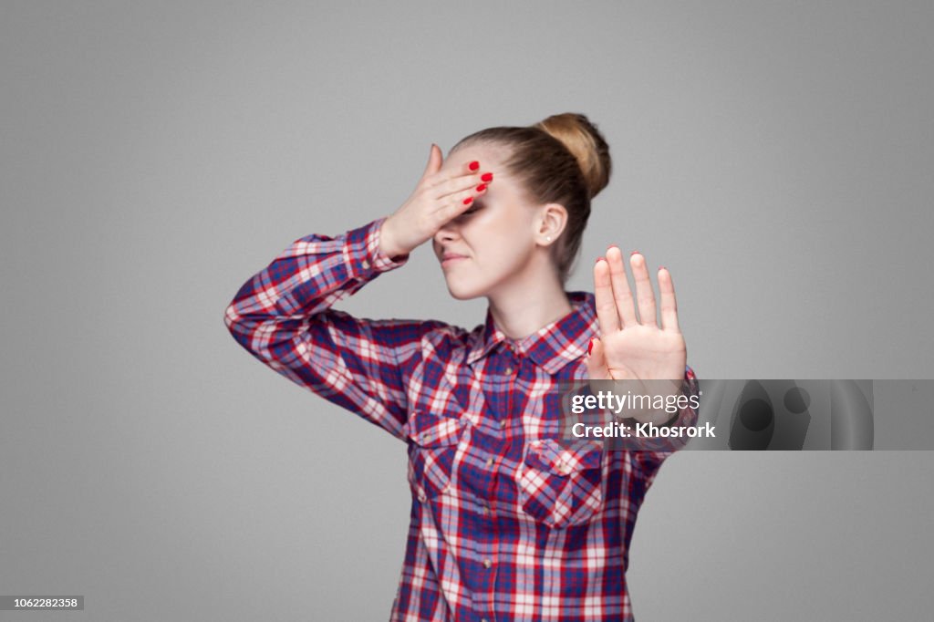 Stop, I don't want to see it. beautiful blonde girl in pink checkered shirt, collected bun hairstyle standing, covering eyes and blocking with hands.