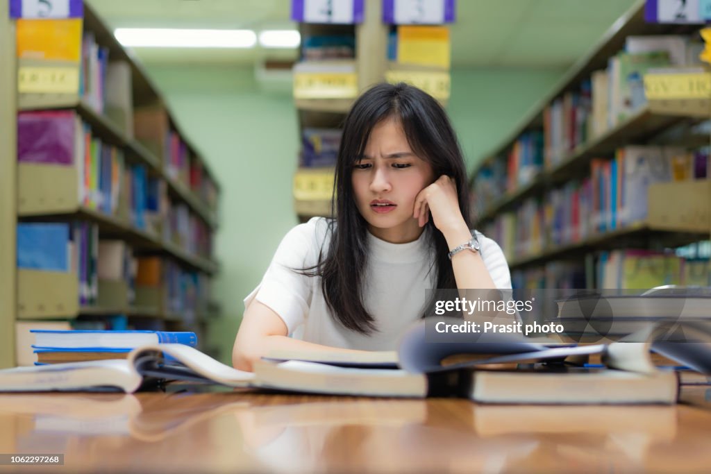 Asian woman student boring reading book at library with a lot of books in university. Asian student disheartened reading book for examination