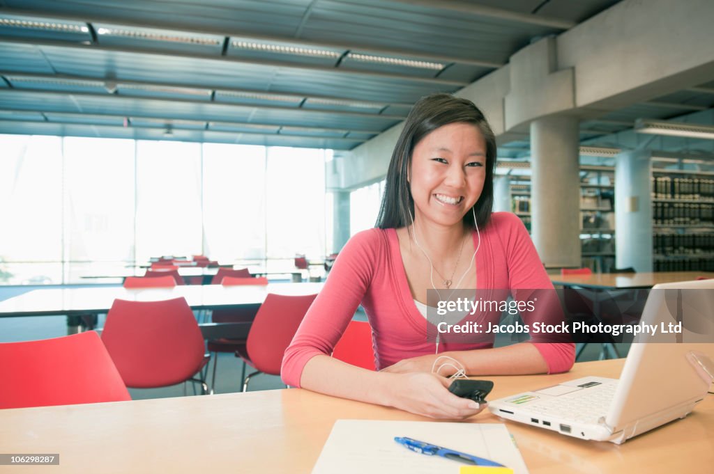 Smiling Chinese student listening to headphones and working at laptop in library