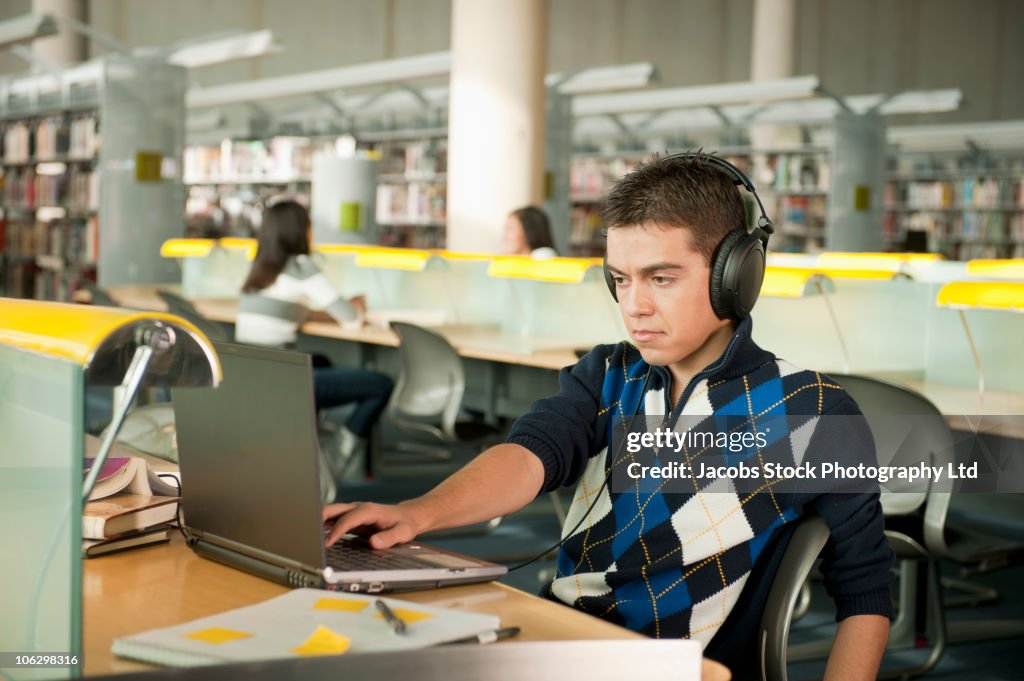 Hispanic student listening to headphones and working at laptop in library