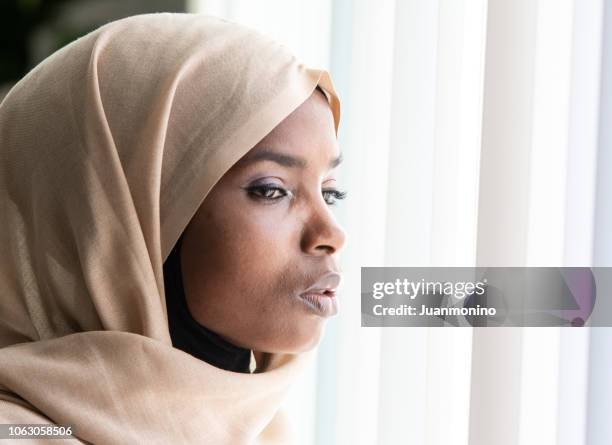 concerned black muslim woman looking through a window - african refugee stock pictures, royalty-free photos & images