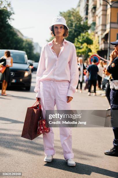 Model Giedre Dukauskaite wears a white Lacoste bucket hat, light pink button-up shirt, light pink striped linen pants, a red print bag, and white...