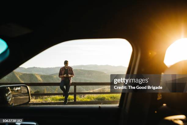 man leaning on fence using mobile phone on country road - telefonnummer wählen stock-fotos und bilder