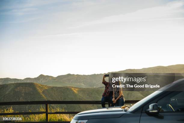 couple taking selfie while sitting on fence in countryside - mobile phone and adventure stockfoto's en -beelden