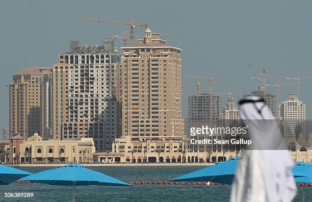 Man wearing traditional Qatari clothing walks along a beachfront opposite new highrise buildings under construction on the man-made peninsula called...