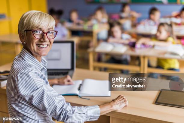maestra de primaria feliz en su escritorio en el aula. - teacher desk fotografías e imágenes de stock