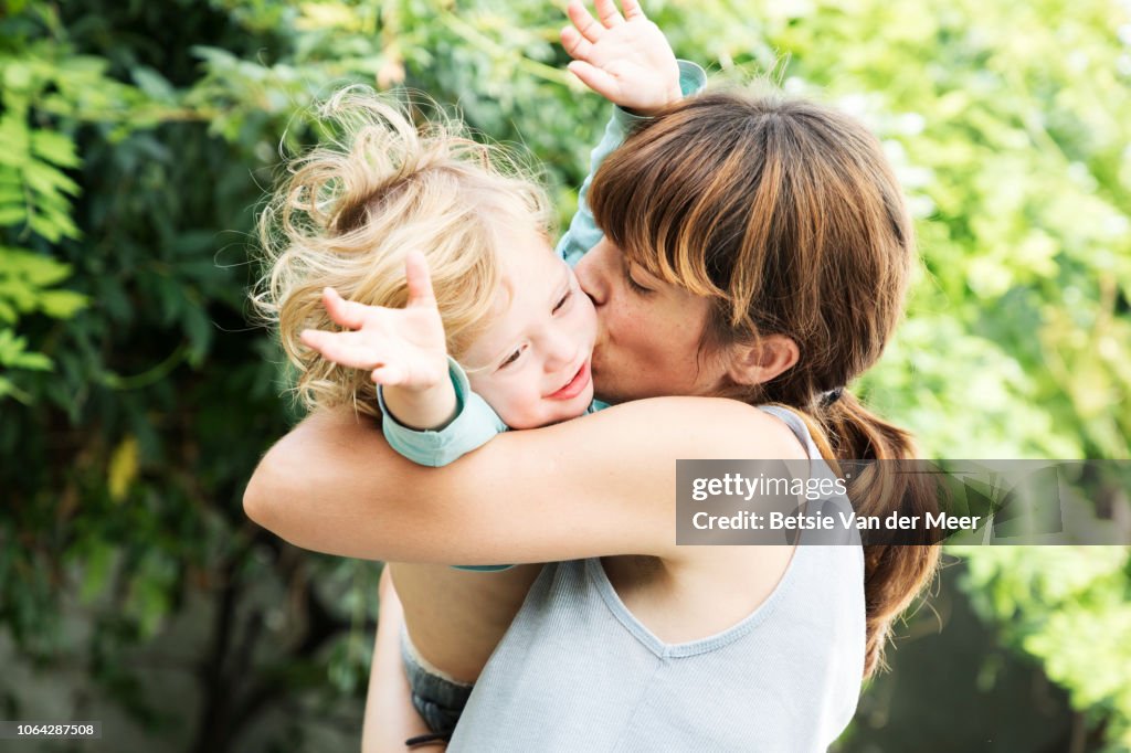 Mother cuddles and kisses toddler in garden.
