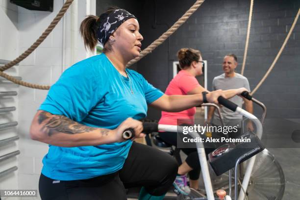 two attractive body-positive women, latino and caucasian, doing a workout on the exercise bike in the gym under the supervision of the coach, the senior 55-years-old cuban hispanic man - alex potemkin or krakozawr latino fitness stock pictures, royalty-free photos & images