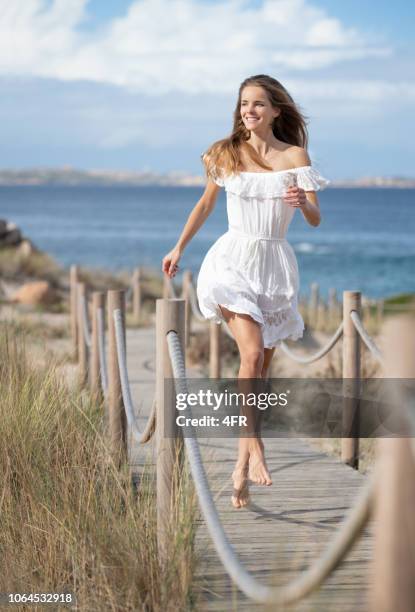 beautiful woman in a white dress with a perfect candid expression skipping down a roped footpath to the beach, sardinia, italy - beach dress stock pictures, royalty-free photos & images