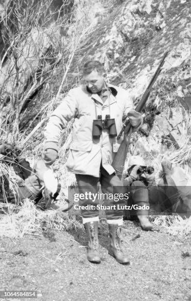 Black and white photograph of a middle-aged man standing in full-length view, wearing a hunting jacket, tall lace-up boots, and mittens, with...