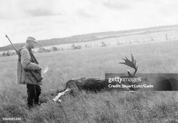 Black and white photograph, shot on an angle, of a middle-aged man, standing in full-length profile view, wearing a hunting jacket and cap, with a...