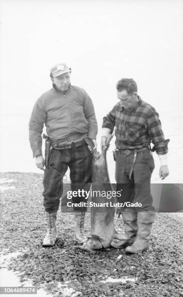 Black and white photograph of two middle-aged men, one wearing a jacket, cap, lace-up boots, a holstered handgun and ammunition at his waist, the...