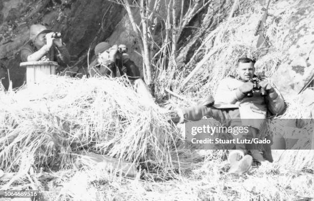 Black and white photograph of three middle-aged men in an outdoor setting with dry grasses and branches on the ground and a rocky hill or cliff in...