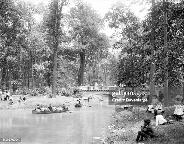 Young children and adults enjoy the quiet of Belle Isle Park in Detroit, MI, 1903. Boaters are visible on the pond, while other park goers stand on a...