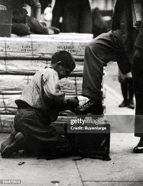 Young Bowery bootblack grimaces with the effort as he kneels and shines the shoes of a passerby in downtown New York, July 1910.