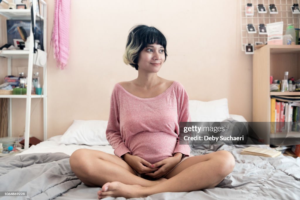 Pregnant brown skin asian woman with short highlighted hair sitting on the bed at room smiling positively
